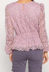 Blusa in pizzo di un delicato rosa con maniche a sbuffo lunghe, caratterizzata da un punto vita peplum, orlo a scallop e una piccola apertura a chiave sul retro.