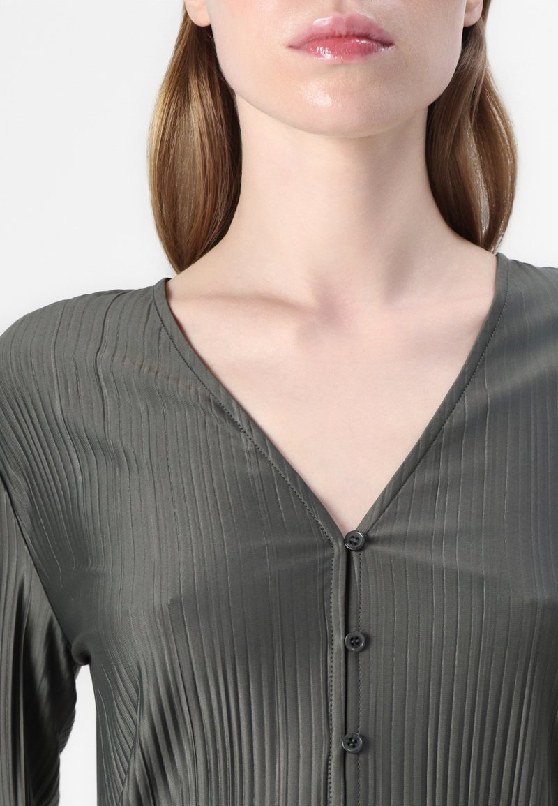 Olivgrünes Shirt mit V-Ausschnitt, mit vertikalen Falten und fünf schwarzen Knöpfen vorne. Glattes, leichtes Material mit einem Schimmer.