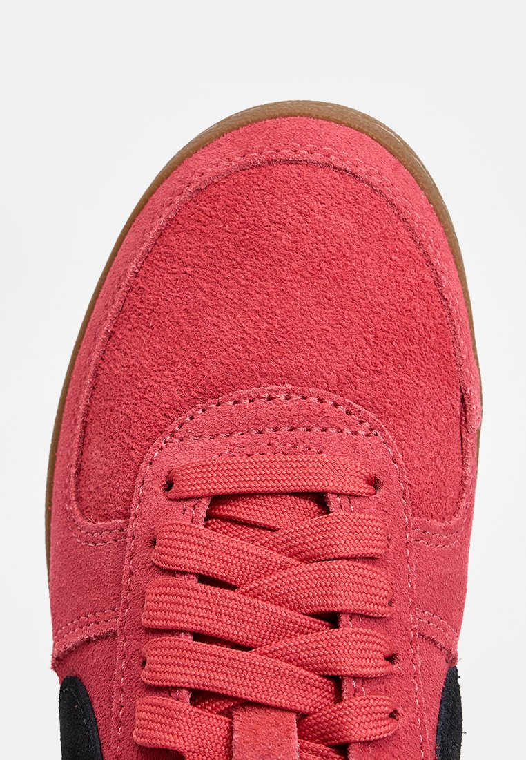 Zapatilla de ante roja con detalles en negro, que presenta una puntera redondeada, cordones trenzados y una superficie texturizada. La suela de goma es de color tostado.