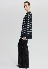 Kvinna står i en långärmad topp med svartvita ränder och svarta paljettbyxor med vida ben mot en enkel ljus bakgrund.