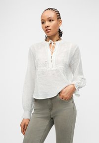 Γυναίκα με πλεξούδες, φορούν λευκό μπλουζάκι με μακριά μανίκια, υφή και δαντελένιες λεπτομέρειες, και γκρι εφαρμοστά παντελόνια, με το χέρι στην τσέπη, σε λευκό φόντο.