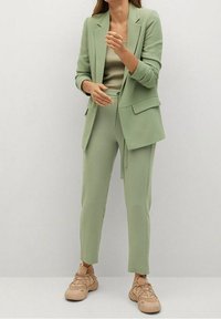 Un costume vert clair composé d'un blazer ajusté avec des poches et d'un pantalon fuselé, associé à un haut beige côtelé et à des baskets claires.