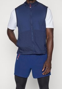 Marineblauer leichter Weste mit hohem Kragen, Frontreißverschluss und roten Hardwareelementen, kombiniert mit strukturierten blauen Shorts im taillierten Schnitt.