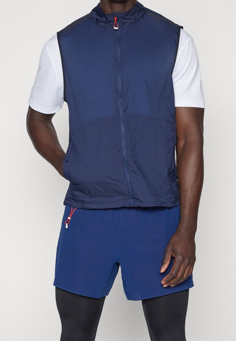 Marineblauer leichter Weste mit hohem Kragen, Frontreißverschluss und roten Hardwareelementen, kombiniert mit strukturierten blauen Shorts im taillierten Schnitt.