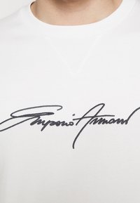 Vit bomull t-shirt med en svart broderad signaturdesign över bröstet, med rund halsringning och en subtil v-formad accent.