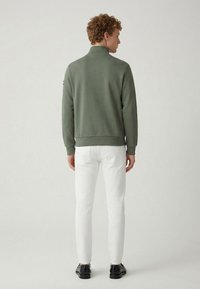 Vihreä collegetakki, jossa on kaulus, resorit hihansuissa ja helmassa, yhdistettynä valkoisiin kapeisiin housuihin ja mustiin kenkiin, kuvattuna takaa.