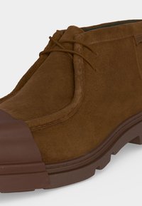 Bottine en daim marron avec fermeture à lacets, semelle en caoutchouc texturée et embout renforcé marron foncé.