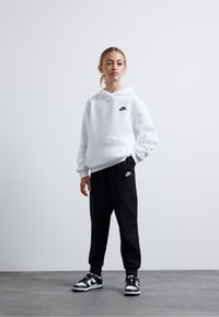 Hanorac alb cu buzunar frontal și logo Nike negru, purtat împreună cu pantaloni de trening negri cu un logo, și adidași negri cu alb.