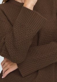 Brauner Strickpullover mit strukturiertem Muster und gerippten Bündchen. Das Material scheint weich und gemütlich zu sein.