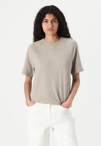 Beige short-sleeve knit t-shirt with a round neckline and raglan sleeves, displayed with hands in pockets of white trousers.