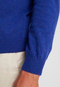 Blå ulltröja med ribbat manschett, som har en mjuk textur och en något lös passform. Synlig sömdetalj på sidan.