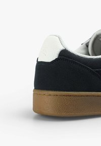 Zapatilla de ante negra con un talón de cuero blanco, suela de goma texturizada marrón y un logotipo sutil en el talón. Forma redondeada, diseño casual.