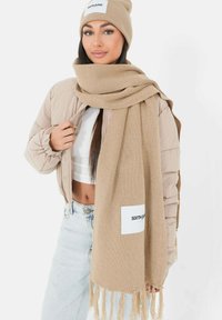 Beige gebreide sjaal met franjes en een wit etiket, gedragen met een beige pufferjack en een witte top, met lichtblauwe jeans.