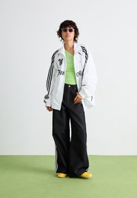 Witte oversized jas met zwarte strepen, groene aansluitende tanktop met een hartontwerp, brede zwarte jeans en gele instappers.
