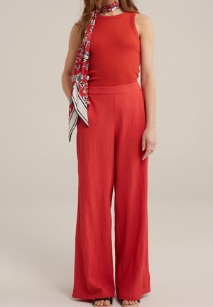 Vrouw met een rood mouwloos topje, losse gestructureerde rode broek en een rood bloemen sjaaltje om haar nek gebonden, staand met één hand langs haar zij.