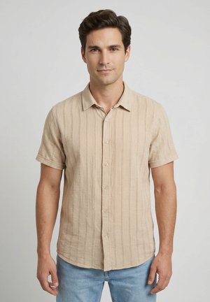 Hombre de cabello oscuro que lleva una camisa beige de manga corta con botones y vaqueros azul claro, posando frente a un fondo liso y claro.