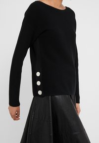 Femme portant un pull noir à côtes avec trois grands boutons en argent sur le côté, associé à une jupe en cuir noire.