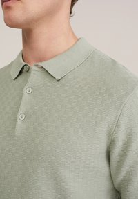 Man draagt een lichtgroen, gestructureerd poloshirt met drie knopen, kraag zichtbaar, close-up van nek tot schouder.
