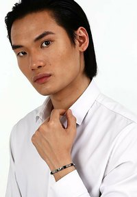 Svart och silverpärlad armband bärs på en handled, med släta och strukturerade pärlor, mot en vit knapp-up skjorta.