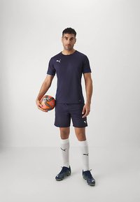 Mees, kes seisab tumesinises spordiriietuses ja valgetes sokkides, hoides vasaku käega oranži-hallikat jalgpalli tavalise tausta ees.