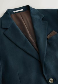 Blazer bleu sarcelle foncé avec revers à cran, doublure intérieure marron, pochette et un bouton beige sur le devant.