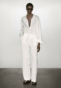 Chemise en lin blanche à col en V profond et manches longues, assortie à un pantalon blanc ample, portée avec des sandales noires. Textures du tissu visibles.