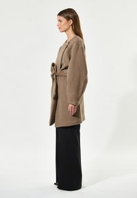 Cappotto di lana marrone con maniche oversize, cintato in vita. Abbinato a pantaloni neri a gamba larga. Design semplice, minimalismo negli adornamenti.