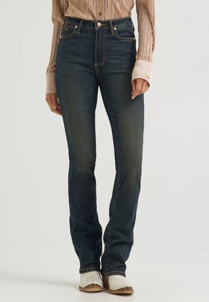 Kvinde iført mørkeblå jeans med lige ben, en skinnende beige langærmet bluse og hvide ankelstøvler, stående mod en ensfarvet baggrund.