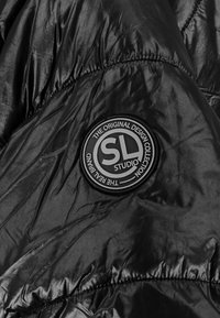 Zwarte pufferjack met een glanzend synthetisch materiaal, een gequiltde textuur en een ronde logo patch met de tekst "SL STUDIO."