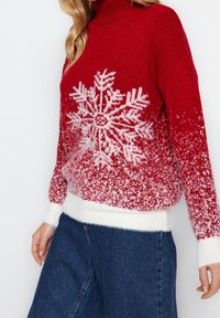 Punainen neule, jossa on valkoinen lumihiutalekuvio, varustettuna korkeakauluksella ja gradienttisuunnittelulla, joka haalistuu valkoiseksi helman ja hihansuiden kohdalla.