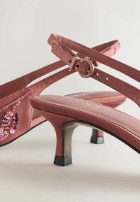 Vaaleanpunaiset satiiniset korkokengät, joissa on ristivarsit, yhdellä sivulla on paljettikoristelu ja ne ovat näyttävän kiiltävät.