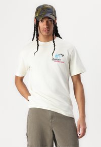 Hvit bomulls T-skjorte med et grafisk design med blå og rosa tekst. iført olivengrønne shorts og en kamuflasje caps. Lange flettehår synlige.