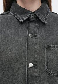 Črna denim srajca z klasičnim ovratnikom, srebrnimi zaponkami in prsnim žepom z okrepljenim robom. Gladek tekstura z rahlimi vzorci.