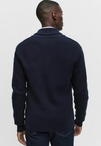 Marinblå ribbad stickad tröja med sjalkrage och långa ärmar. Har en figurnära nederkant och en strukturerad mönster. Visad bakifrån.