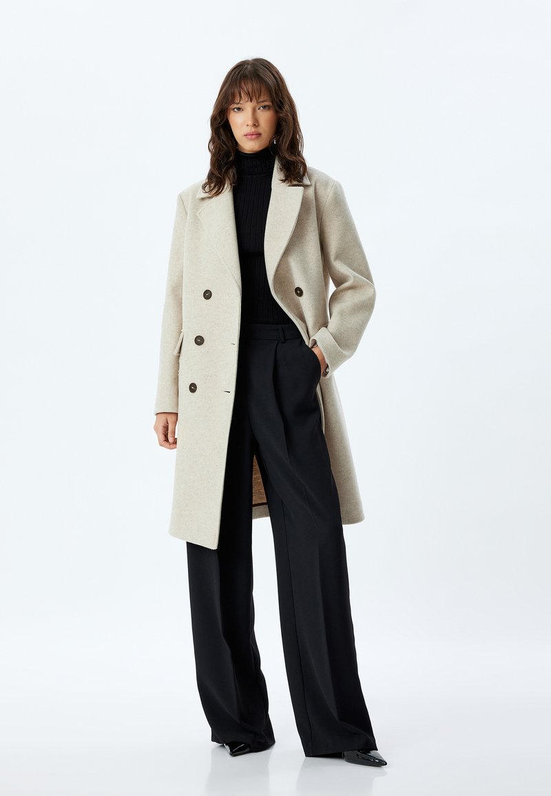 Beige uldblanding dobbeltbrystet frakke med runde revers, brune knapper og sidelommer, parret med sort turtleneck og brede bukser.