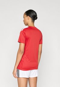 Κόκκινη αθλητική φανέλα με κοντά μανίκια και στρογγυλή λαιμόκοψη, με λευκές ρίγες στους ώμους. Συνδυάζεται με λευκό σορτς και μαύρες λεπτομέρειες.