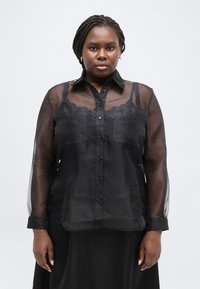 Γυναίκα με πλεξούδες, που φορά χρυσά σκουλαρίκια και διαφανές μαύρο πουκάμισο με κουμπιά πάνω από μαύρο τοπ, στέκεται μπροστά σε ένα απλό φόντο.