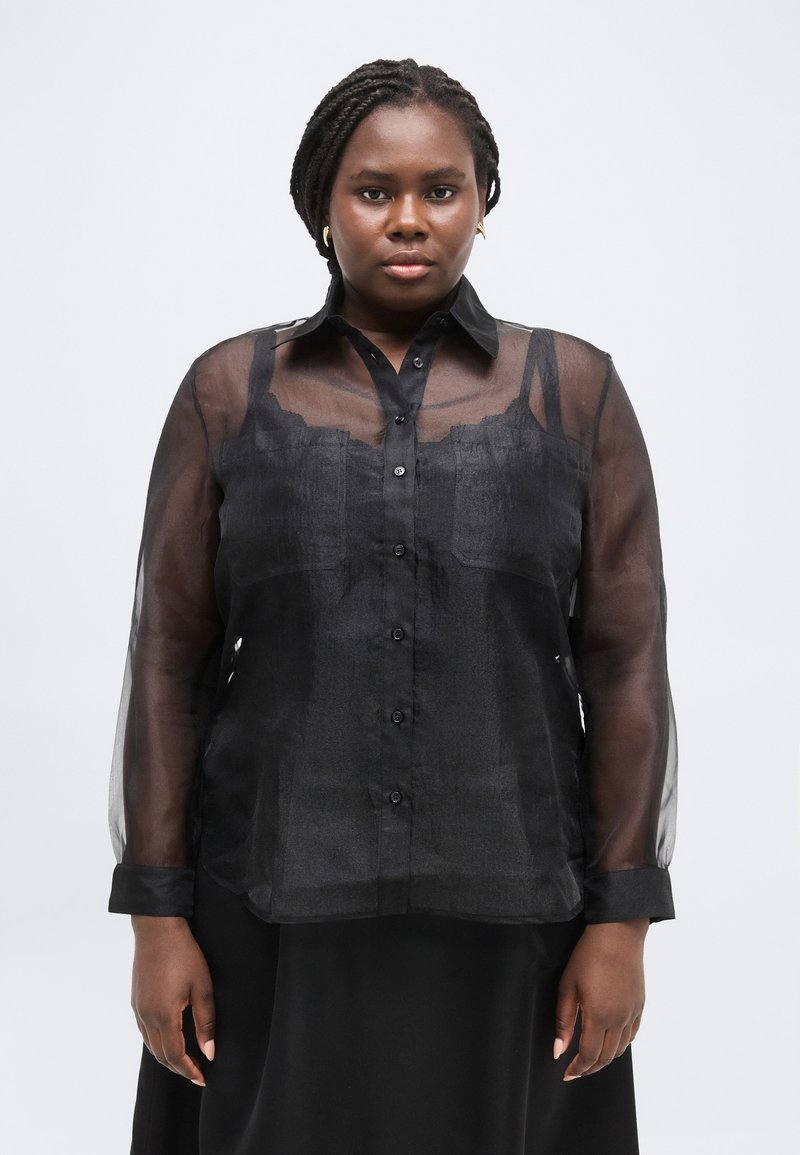 Γυναίκα με πλεξούδες, που φορά χρυσά σκουλαρίκια και διαφανές μαύρο πουκάμισο με κουμπιά πάνω από μαύρο τοπ, στέκεται μπροστά σε ένα απλό φόντο.