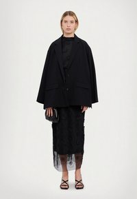 Melns plaša piegriezuma žakete ar asām apļveida atlokām, pārsvilkts pāri garai melnai kleitai ar ziedu mežģīņu rakstiem un caurspīdīgu apakšmalu, papildināts ar nelielu rokassomu.