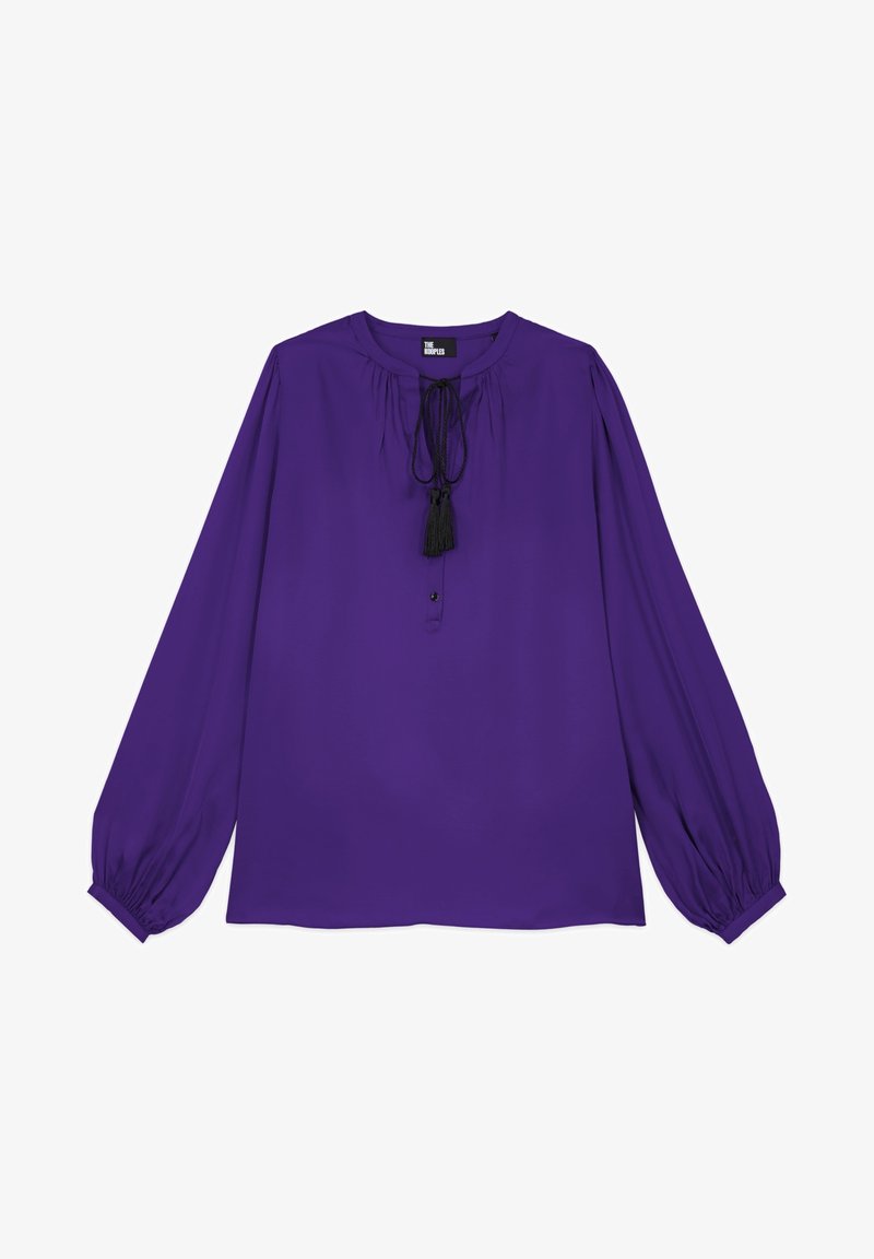 Blouse violette à manches longues avec poignets froncés, encolure ronde et lien noir à pompons au centre devant.