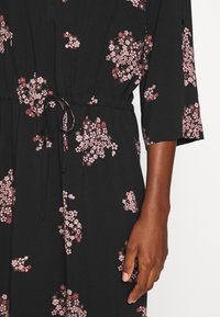 Musta kukkakuosiset mekko, jossa on vaaleanpunaisia ja punaisia kuvioita. Varustettu säädettävällä nauhalla vyötäröllä ja kolmen neljänneksen hihoilla. Suunniteltu sileästä kankaasta.