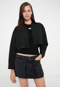 Zwarte cropped jack van gladde stof met een ritssluiting, gecombineerd met een aansluitend zwart ribbeltop. Zwarte spijkerrok met zijzakken.