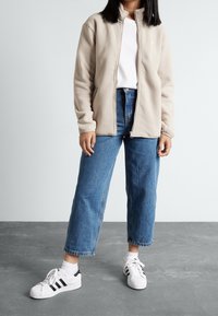 Chaqueta de forro polar beige con cuello alzado y cierre de cremallera, combinada con una camiseta blanca y unos vaqueros de cintura alta azules, junto con zapatillas blancas.