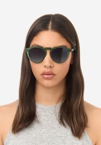 Žalios spalvos akiniai nuo saulės su apvaliais rėmeliais, tamsių gradientinių stiklų ir unikaliu tiltelio dizainu. Lengvos medžiagos ir lygi tekstūra.