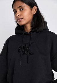 Felpa nera con cappuccio realizzata in tessuto morbido e dalla vestibilità oversize. Presenta un logo "CK" nero in rilievo e un cappuccio regolabile con cordino.
