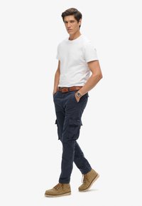 Camiseta blanca de algodón, pantalones cargo azul marino con múltiples bolsillos, cinturón marrón y botas color arena. El modelo se mantiene de pie con confianza con las manos en los bolsillos.