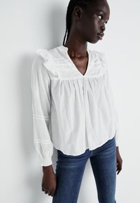 White blouse with ruffled collar and pleated details. Long sleeves with gathered cuffs. Paired with fitted blue jeans.