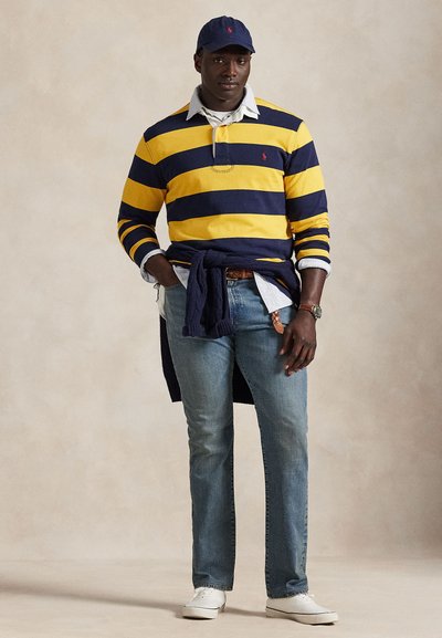 Mand iført navyblå og gule stribede langærmede skjorte, lyseblå jeans, hvide sneakers, navy kasket og sweater bundet om taljen.