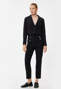 Blazer nero accorciato con colletto a scollo a V e chiusura con un bottone, abbinato a pantaloni neri su misura con doppi bottoni dorati.