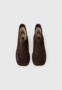 Botine din suede maro la glezna, cu un căptușeală moale din blană, margini rotunjite și talpă plată, având un design simplu și minimalist, fără accesorii metalice.
